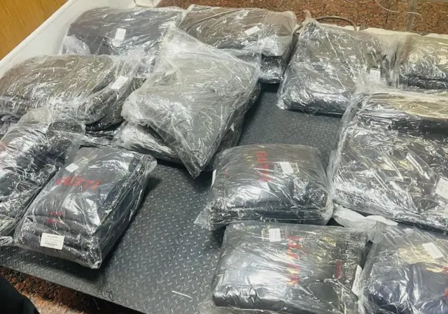 Several sealed black bags wrapped in clear plastic lay on a textured metal surface, stacked haphazardly in a workspace.