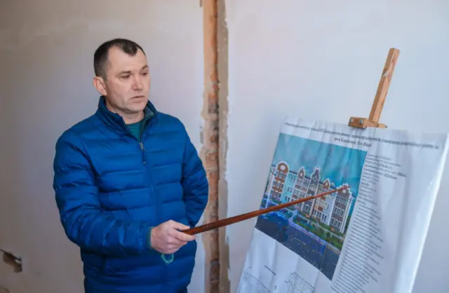 Man in a blue jacket points to an architectural rendering on a poster attached to an easel inside a building under renovation, explaining the plan.