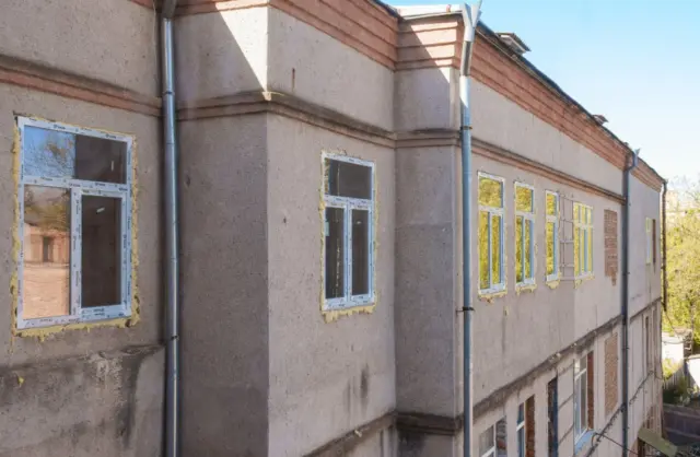 Exterior of a multi-story building under renovation; new window frames wrapped in protective tape along a beige facade with visible downspouts.