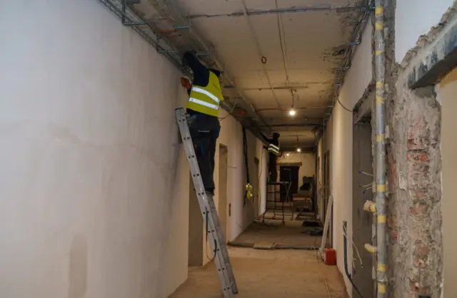 Construction workers in reflective vests on ladders fixing the ceiling in a long hallway under renovation, with exposed wiring overhead.