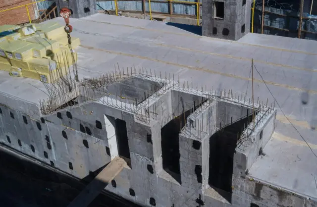 Construction site on a concrete rooftop with stacked insulation, rebar cages, and a hanging crane hook nearby.