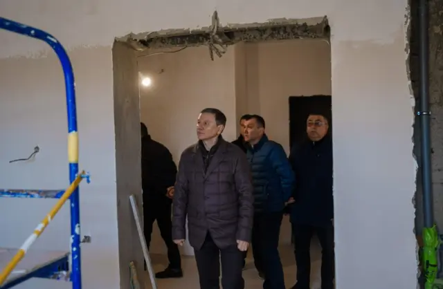 Group of men in winter jackets touring a construction site inside a partially finished room, with blue scaffolding on the left.