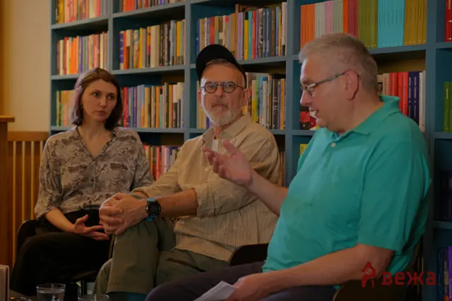 Three people sit in a library discussion; the bearded man in the middle with a cap gestures as the others listen, with colorful books on blue shelves behind them.