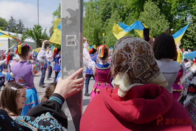 Dancers in colorful traditional Ukrainian costumes perform in a street festival, waving blue-and-yellow flags around a central lamppost.