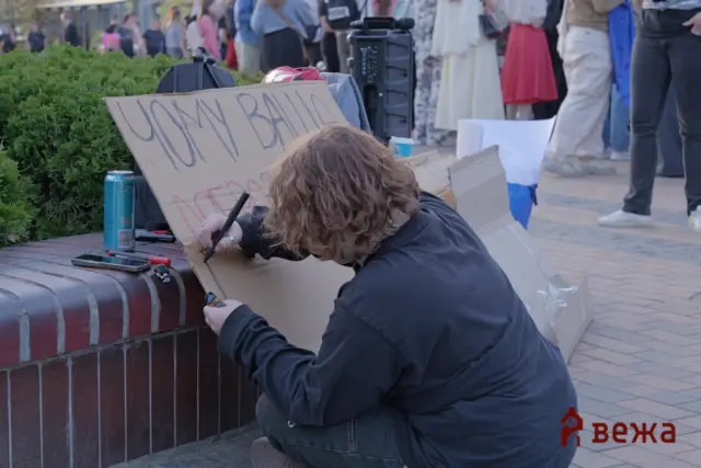 Person kneeling beside a brick planter, drawing on a cardboard sign with a black marker at a crowded outdoor event.