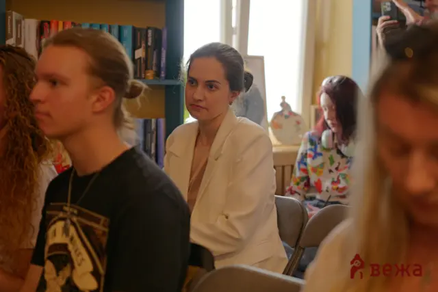 Group of people seated indoors, a woman in a white blazer at center looking thoughtful, others around.