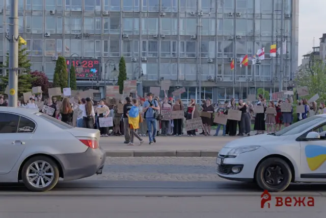 Large crowd of protesters holding handmade signs in a city street, with a glass-fronted building and flags in the background; cars parked along the curb in the foreground.