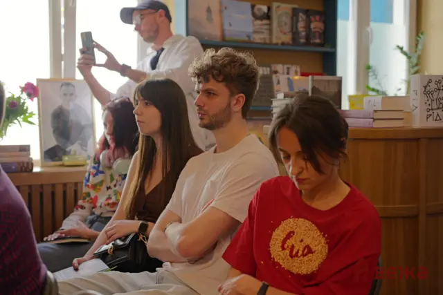 Group of young adults seated in a room with bookshelves and plants; a man in white sits center, a woman beside him, and a person in a red shirt foreground looks down while a man in a cap takes a photo in the background.