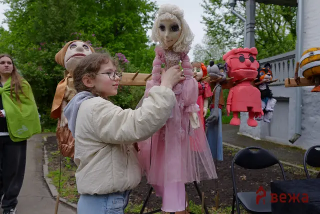 Girl adjusts a pink marionette while colorful puppets hang on a rack behind her outdoors.