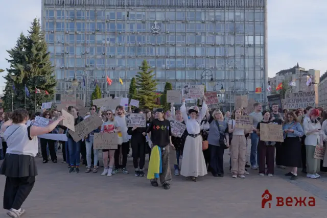 Crowd of protesters holding handwritten placards with Cyrillic text in a city square, in front of a tall glass-fronted building with flags.
