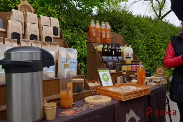 Outdoor market stall with bottles of juice or syrup, jars, and labeled packages on wooden displays, greenery in the background, and a person in red on the right side.