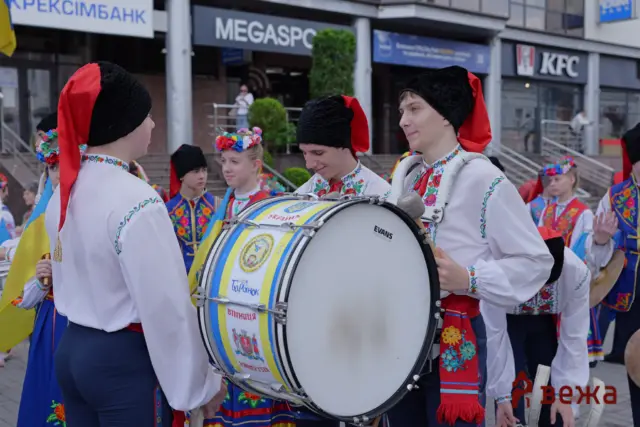 Group of people in colorful Ukrainian folk costumes marching in a street parade, with a large bass drum in the foreground.