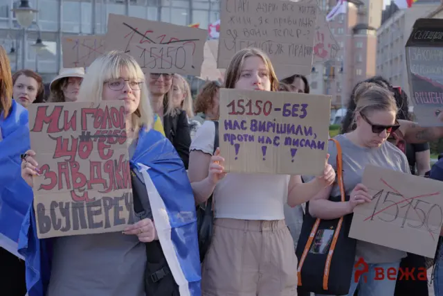 Group of protesters holding handmade cardboard signs with Cyrillic text during a street demonstration in an urban area.