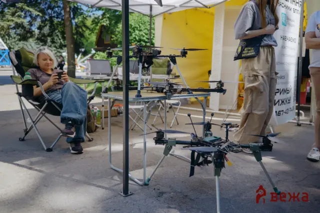 Person relaxing in a camping chair at an outdoor booth, looking at a smartphone, with quadcopter drones on a table nearby.