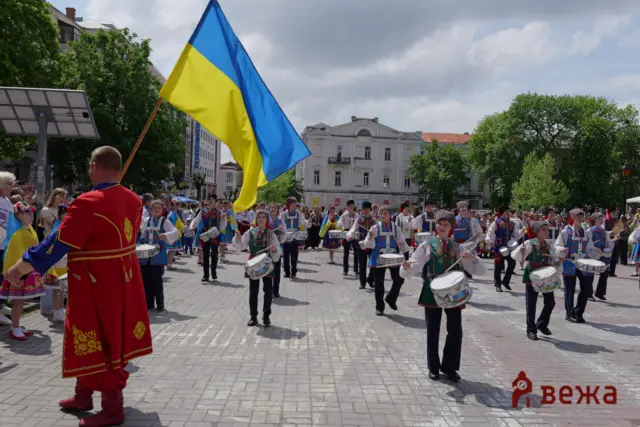 Ukrainian flag waved above a street parade of performers in colorful traditional outfits and drummers in the foreground.
