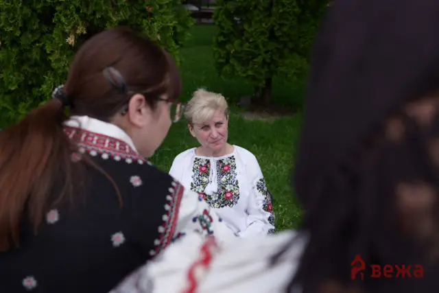 Woman with blond hair wearing a white embroidered blouse speaks to others in a green outdoor setting, seen from the side and foreground figures partially blocking the view