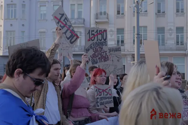 Crowd of protesters holding cardboard signs with large numbers and red crossing lines, standing on a city street with pastel buildings in the background.