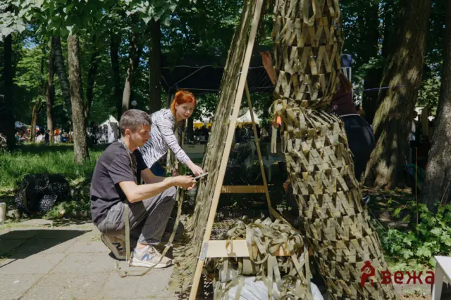Two people crouch beside a tall woven sculpture made of beige strips in a sunny park during a festival.
