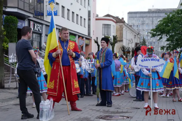 Group of people in bright Ukrainian folk costumes marching in a city parade, one man holding a Ukrainian flag tall flagpole at the front.