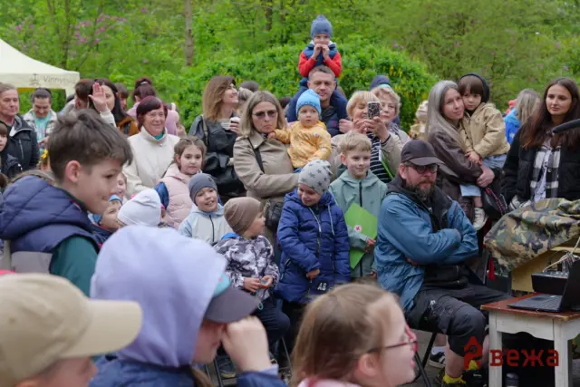 Crowd of families and children at an outdoor park event, some smiling and watching something off-camera. A man holds a child on his shoulders among the group.