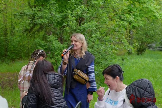 Woman speaks into a handheld microphone to a small crowd outdoors among green trees.