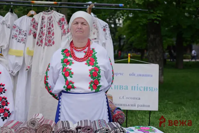 Woman in a white vyshyvanka with red/green floral embroidery and a white headscarf, wearing red bead necklaces at an outdoor market with hanging embroidered garments behind her.