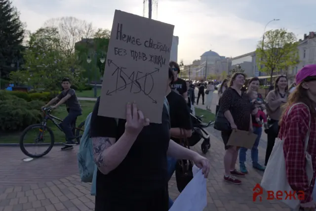 Person in a protest holding a cardboard sign with handwritten Cyrillic text, others march in the background on a city street with trees and bicycles nearby.