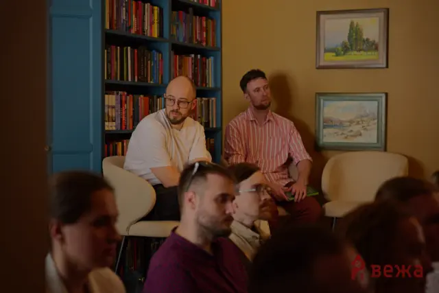 Two men sit on a beige couch in a warm room, with a blue bookshelf and framed landscape paintings on the wall behind them, audience in foreground.