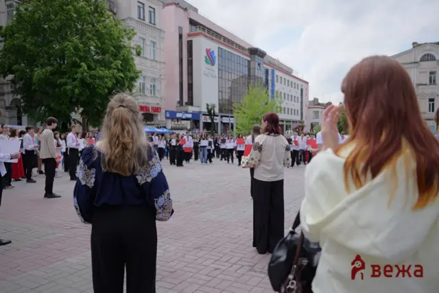People in traditional embroidered clothing form a line in a city square, holding music sheets for a performance while spectators watch.