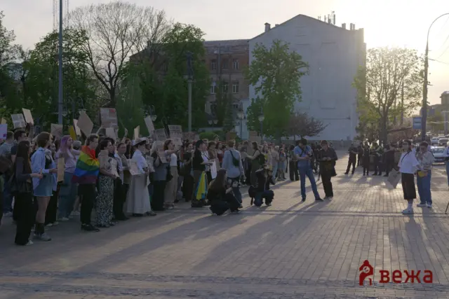 Large group of protesters holding signs in a city square, with a rainbow flag visible on the left side.
