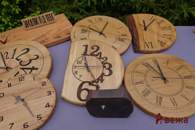 Assorted handmade wooden clocks with Roman numerals laid on a purple surface, with greenery in the background.
