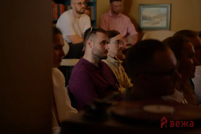 Group of adults seated in a dimly lit room, attentively listening to a presentation with bookshelves and a framed painting in the background.