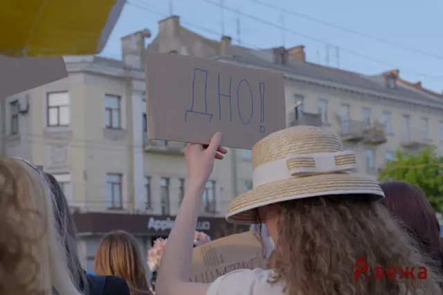 People at a street protest holding up a cardboard sign that reads “NO!” with others and buildings in the background.