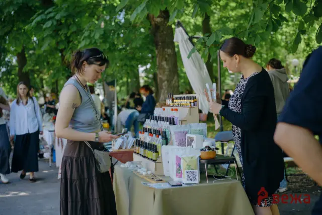 Two women at an outdoor market stall examining skincare products displayed on a table under leafy trees