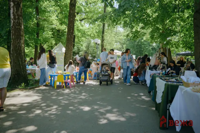 People browsing vendor stalls at an outdoor park market beneath leafy trees.