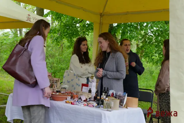 Group of women chatting at an outdoor market stall under a yellow canopy, table filled with jars, bottles, and small goods covered with a white cloth.