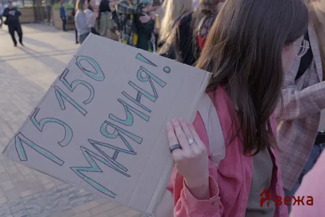 Person with long hair and glasses wearing a pink jacket holds a large cardboard protest sign with blue-green handwritten letters; a crowd stands behind in an outdoor setting.
