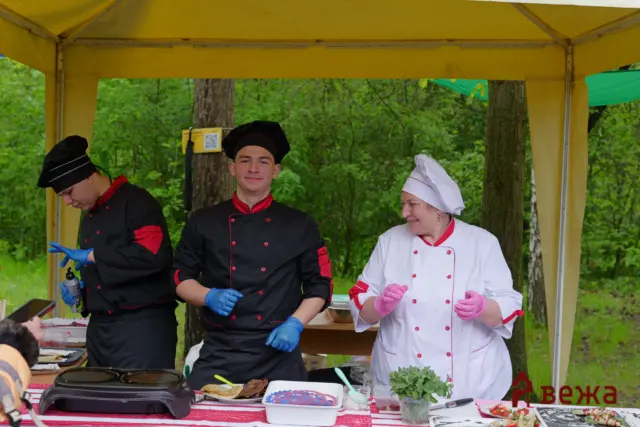 Three chefs under a yellow canopy cooking at an outdoor food stall in a wooded park, smiling at the camera.
