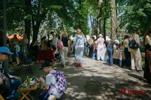 Outdoor park market scene with people browsing vendor tables under leafy trees, children playing with toys in the foreground.