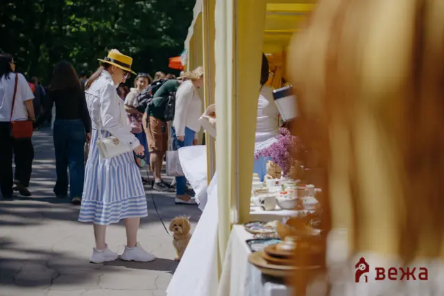 Woman in a striped dress and yellow hat browses a jewelry stall at an outdoor market, small dog on a leash nearby.
