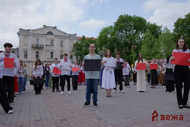 Group of youths in traditional Ukrainian clothing holding colored cards in a city square, with a pale building and green trees behind them.