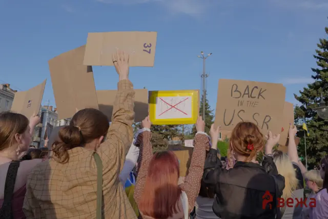 Crowd of protesters holding handmade cardboard signs above their heads against a clear blue sky at a demonstration.