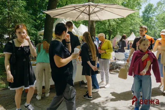 Group of people at an outdoor market or festival under trees, with stalls, tents, and a large beige umbrella overhead.