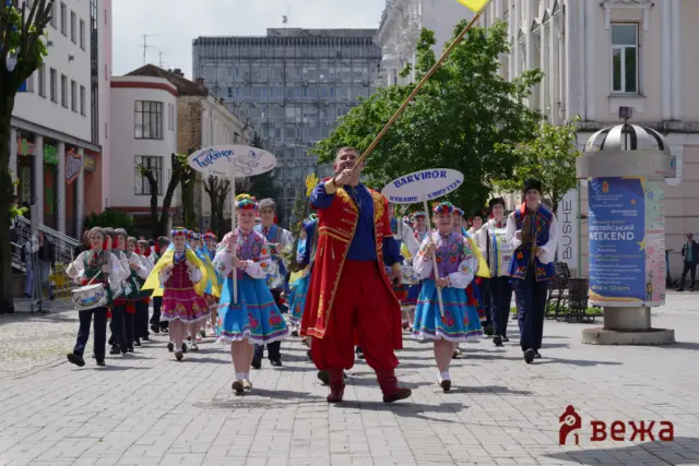 Group of performers in traditional Ukrainian costumes marching in a city street parade, led by a man in bright red attire.