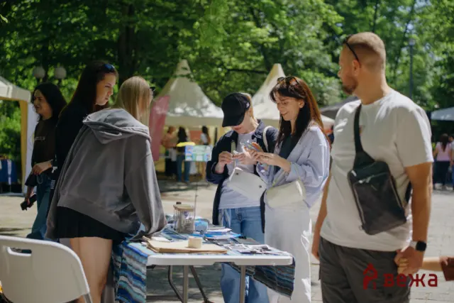 Group of people gathered around an outdoor market table, browsing brochures and items under trees with tents in the background.
