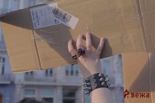 Person wearing a black spiked bracelet holds up a cardboard box with a barcode label outdoors.