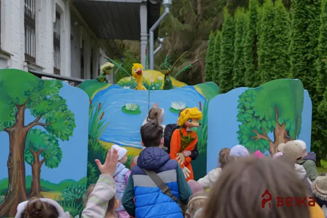 Children watch a colorful outdoor puppet show with bird characters against a painted nature backdrop on stage.
