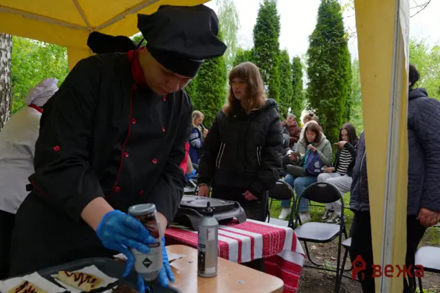 Chef in a black uniform and blue gloves prepares food at an outdoor booth under a yellow canopy, with seated spectators behind him.