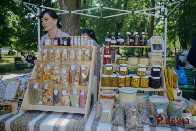 Outdoor market stall in a park with jars, dried goods, and honey on wooden shelves beneath a canopy.