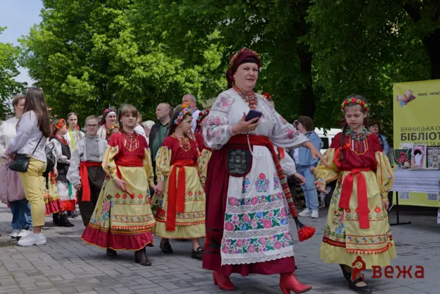 Group of people, mostly children and adults, wearing colorful traditional Ukrainian folk costumes at an outdoor festival under green trees.